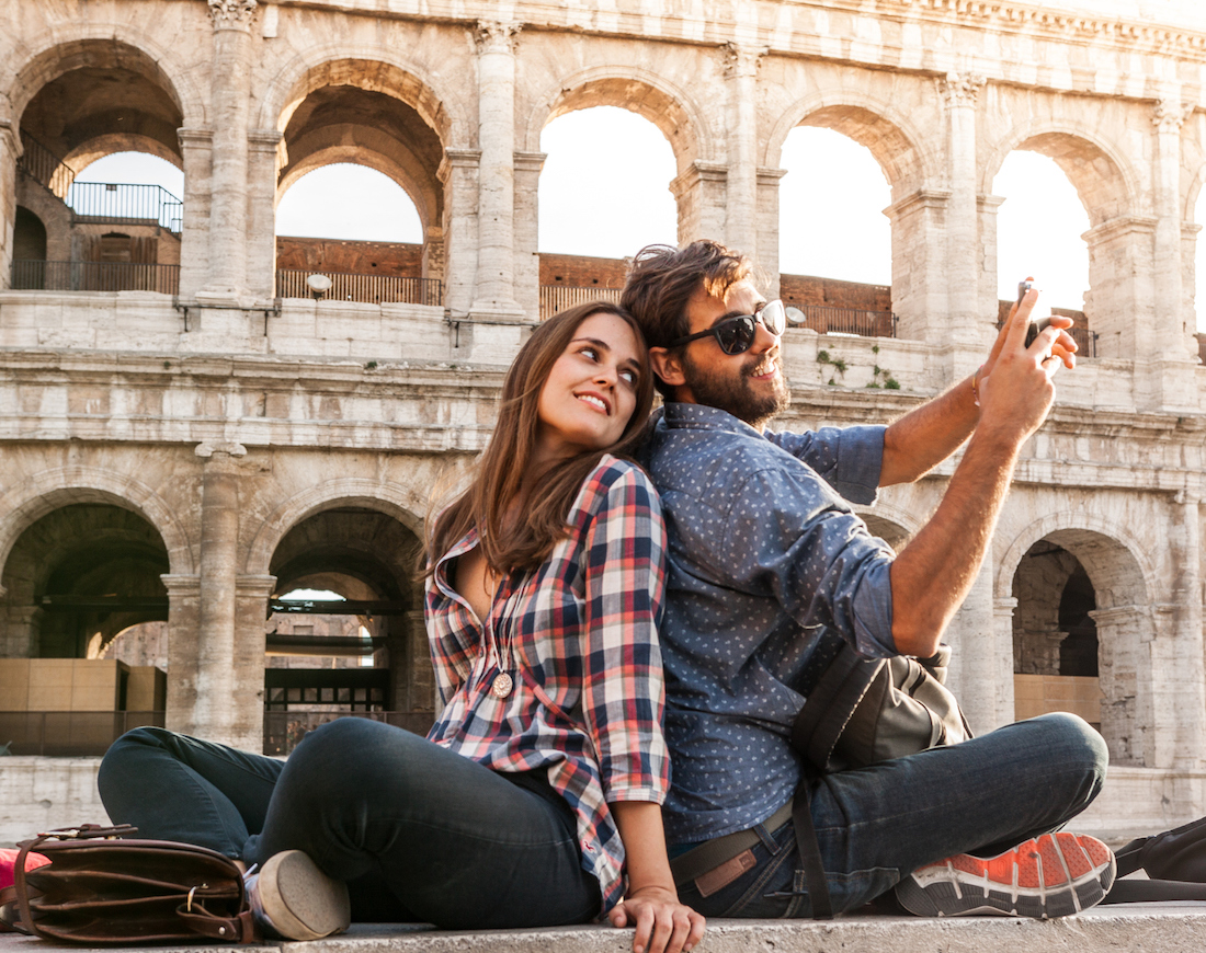 Junges Pärchen sitzt Rücken an Rücken auf dem Boden vor dem Kolosseum in Rom und macht ein Selfie