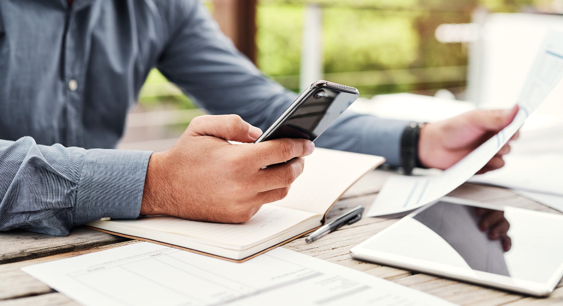 Ein Mann sitzt mit seinem Smartphone, einem Notizbuch, einem Tablet und einigen ausgedruckten Formularen am Tisch