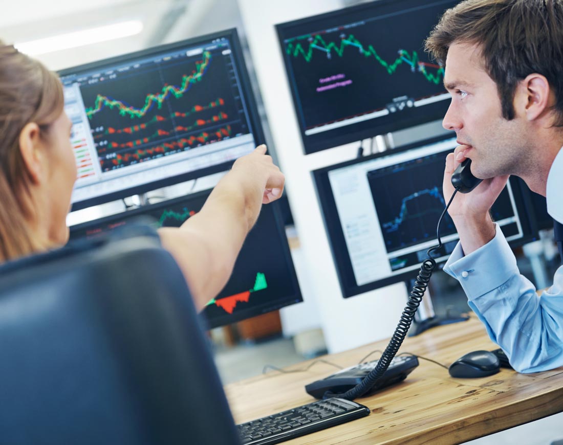Börsenhändler sitzen vor einer Bildschirmwand und beobachten Kursentwicklungen, während sie gleichzeitig telefonieren