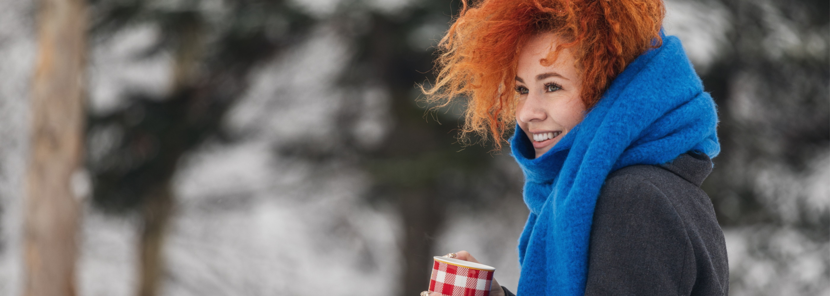 Junge Frau steht lächelnd in einer Winterlandschaft und hält einen Becher in der Hand