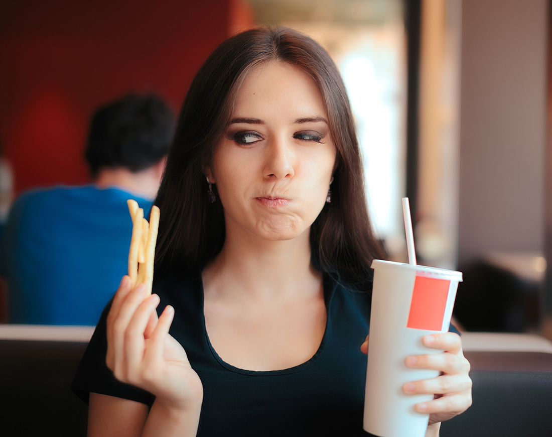Junge Frau sitzt im Fastfood-Restaurant und blickt misstrauisch auf einige Pommes Frites in ihrer Hand