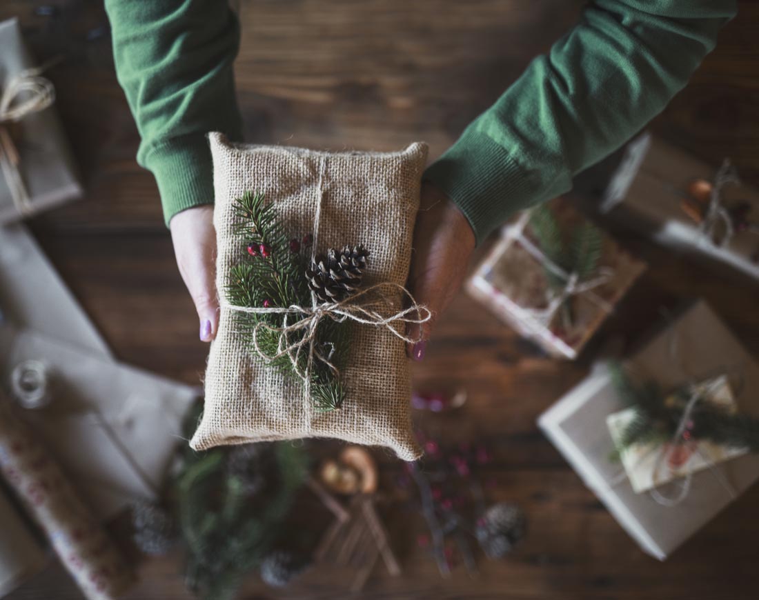 Eine Frau hält über einem Basteltisch eine Geschenkverpackung aus Stoff und mit Tannengrün verziert hoch