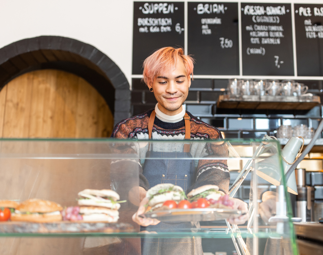 Ein junger Mann mit orange gefärbten Haaren holt einen Teller mit zwei belegten Sandwiches aus Glastheke in einem Café