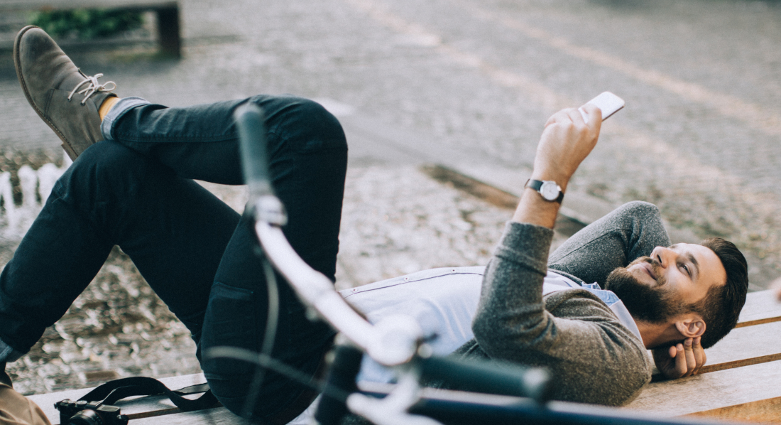 Fahrradfahrer liegt auf einer Straßenbank und blickt lächelnd auf sein Handy