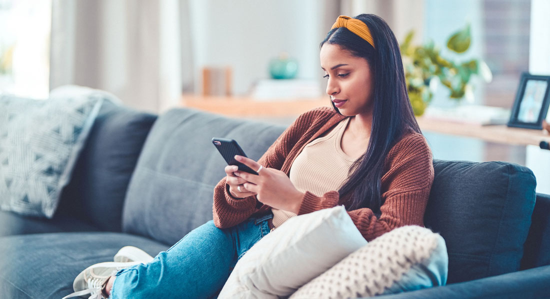 Junge Frau sitzt auf dem Sofa und blickt kritisch auf ihr Smartphone
