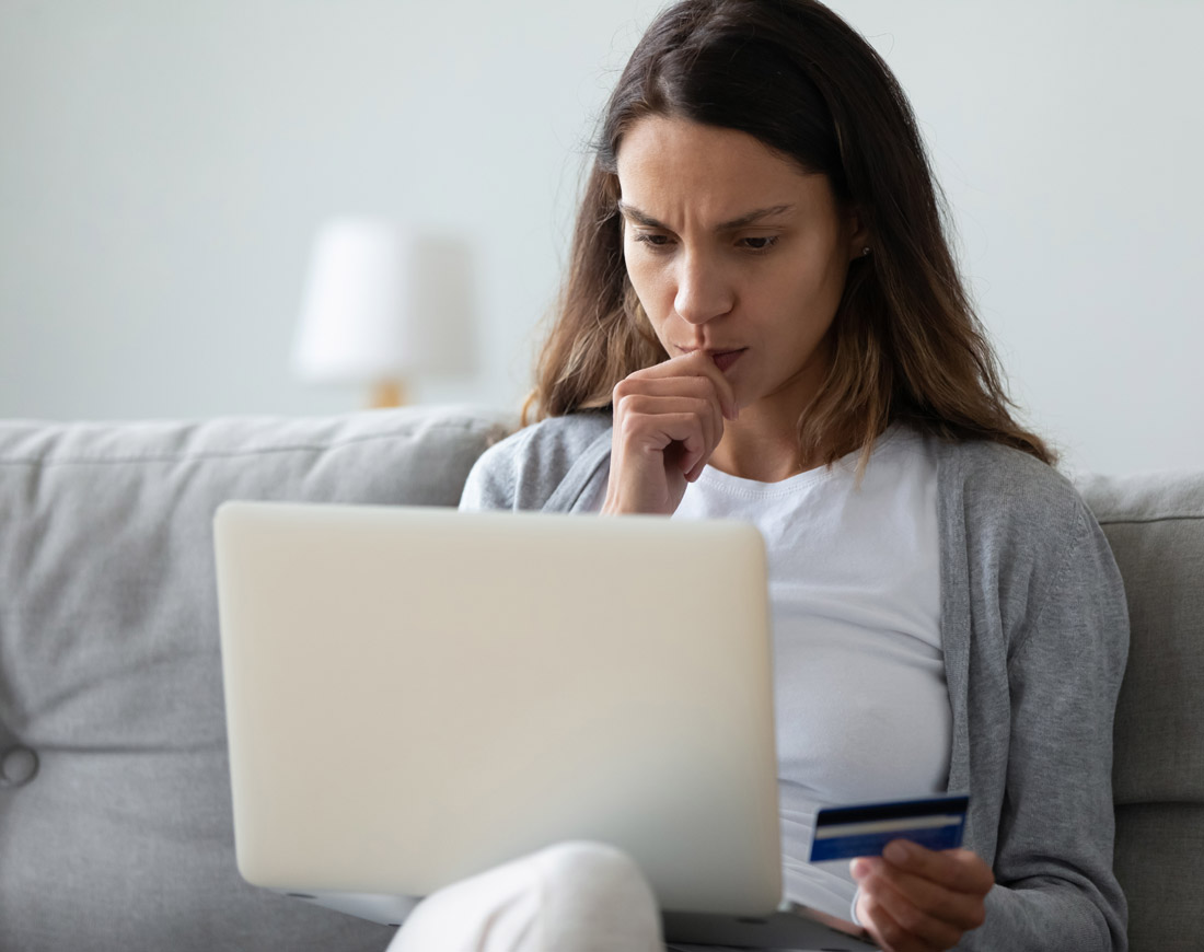 Eine junge Frau sitzt nachdenklich mit einem Laptop auf dem Sofa und hält ihre Kreditkarte in der linken Hand