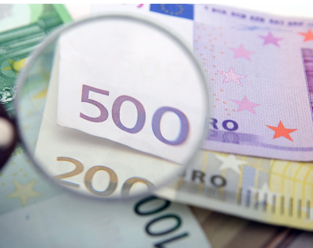 Eine Lupe vergrößert den Wert je eines 500- und 200-Euro-Scheins, die auf einem Tisch zusammen mit einem 100-Euro-Schein liegen