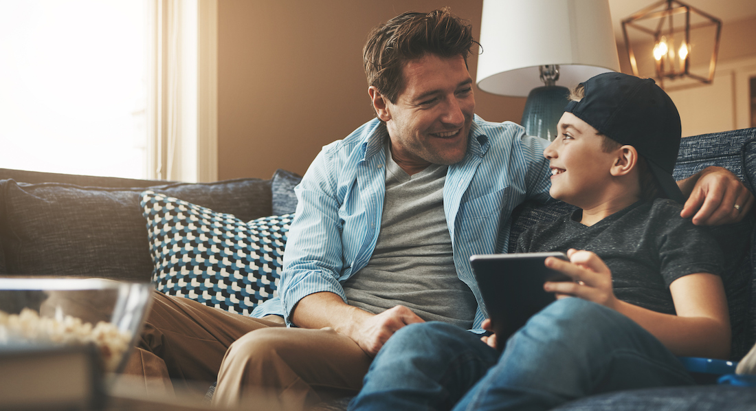 Vater und Sohn sitzen mit Tablet auf einem Sofa und unterhalten sich über Bank-Apps
