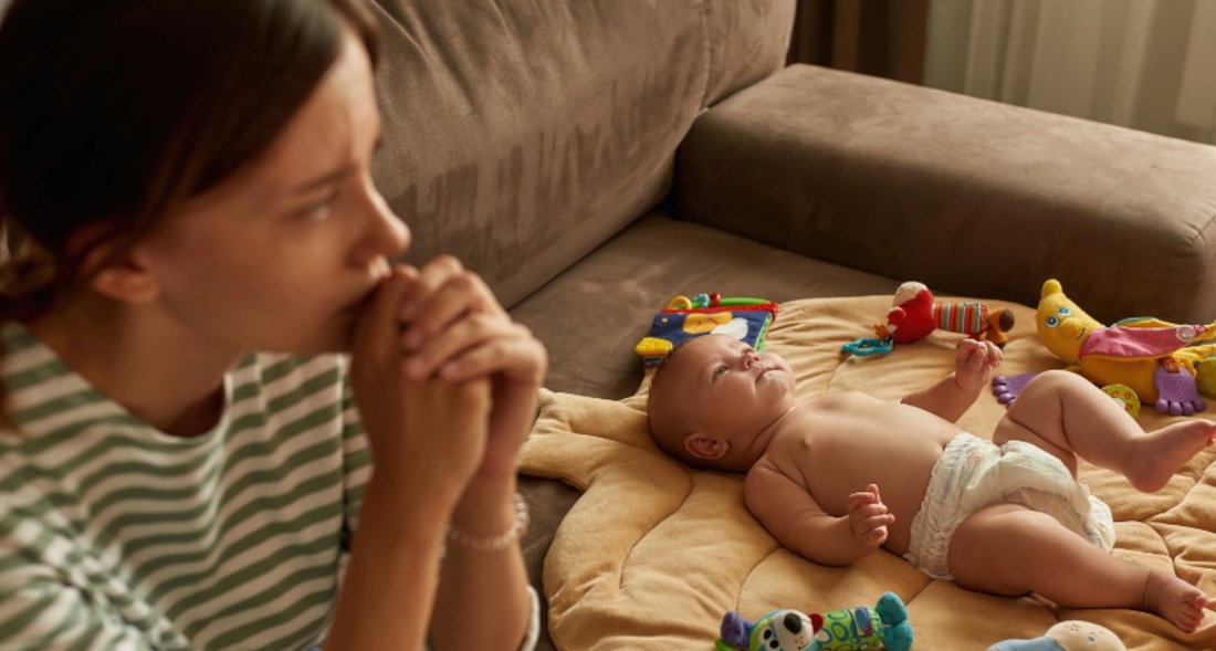 Junge Mutter sitzt nachdenklich auf dem Sofa, ihr Baby auf einer Decke neben sich