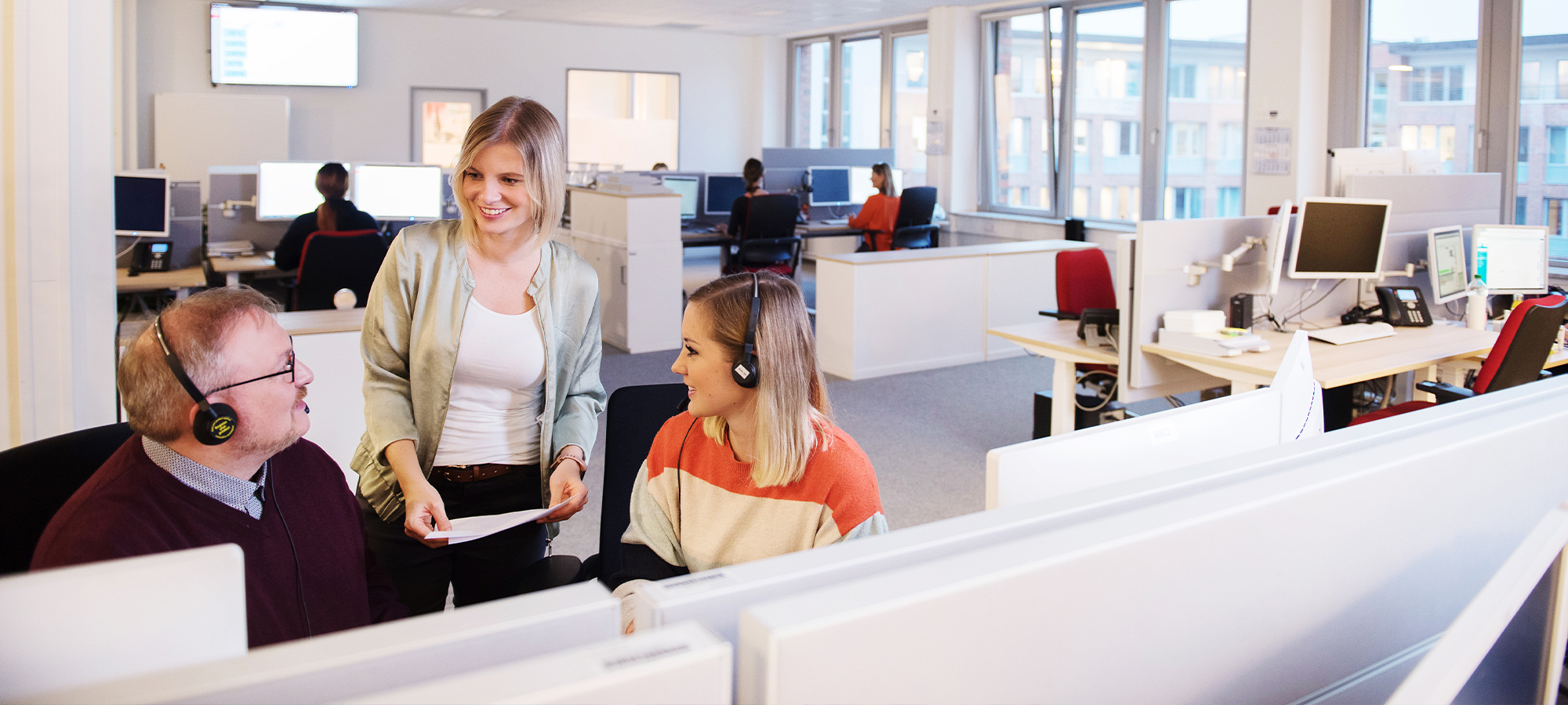 Zwei Personen sitzen vor PC-Monitoren. Sie tragen Headsets. Eine weitere Person steht dabei und spricht mit den beiden.