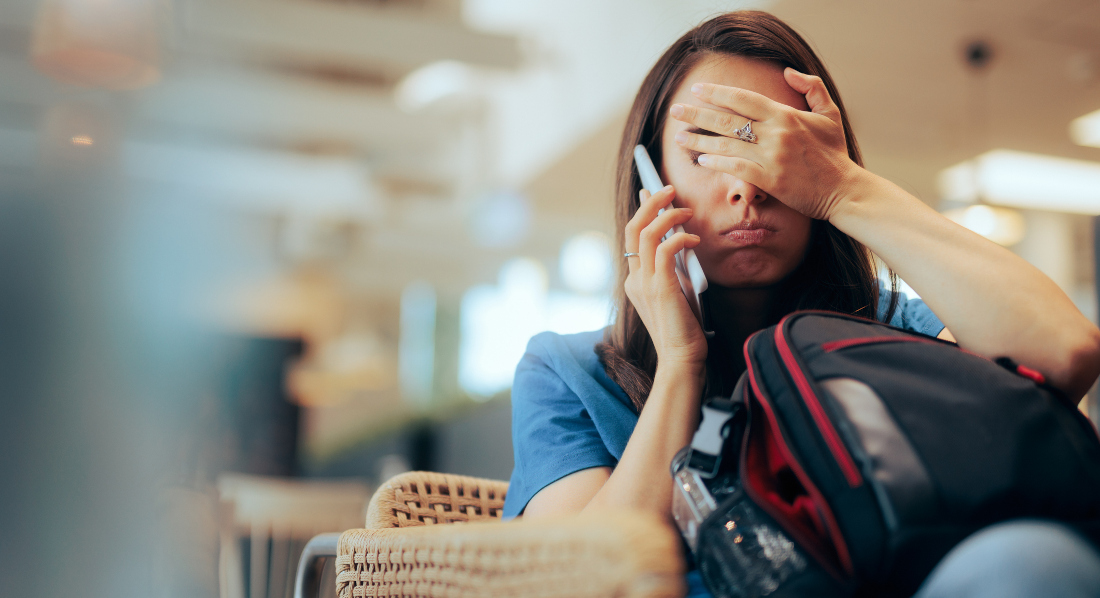 Eine Frau sitzt mit Gepäck am Flughafen. Sie telefoniert und hält eine Hand vors Gesicht. Sie sieht gestresst und unglücklich aus.