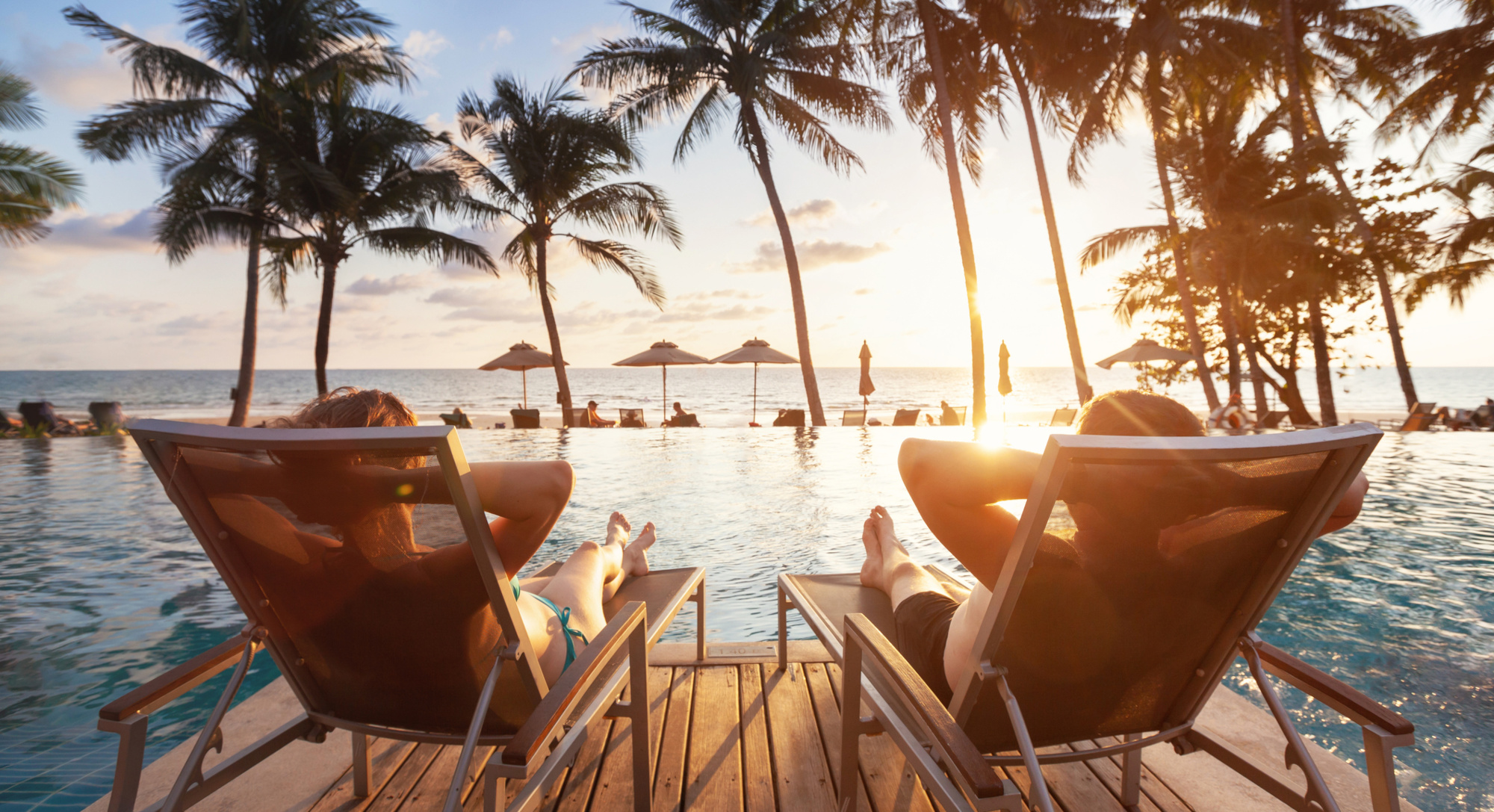 Zwei Personen auf Liegestühlen vor einem Pool und Palmen sehen in den Sonnenuntergang