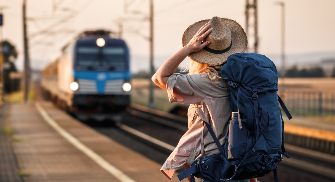 Frau mit Backpacking-Rucksack und Hut steht am Bahngleis und blickt einfahrendem Zug entgegen