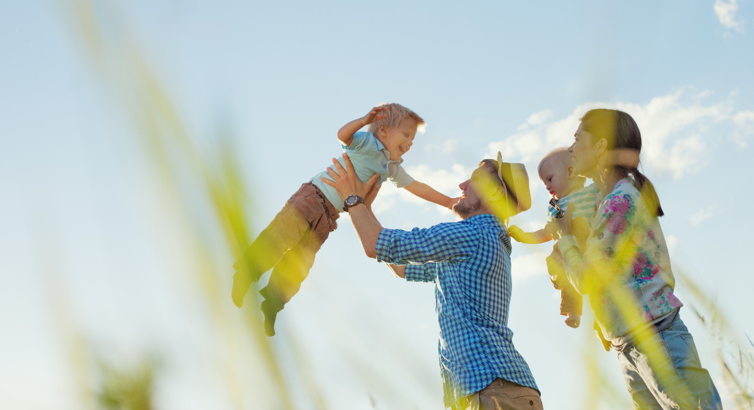 Eltern spielen vor blauem Himmel mit zwei Kindern, der Vater hebt den Sohn hoch in die Luft