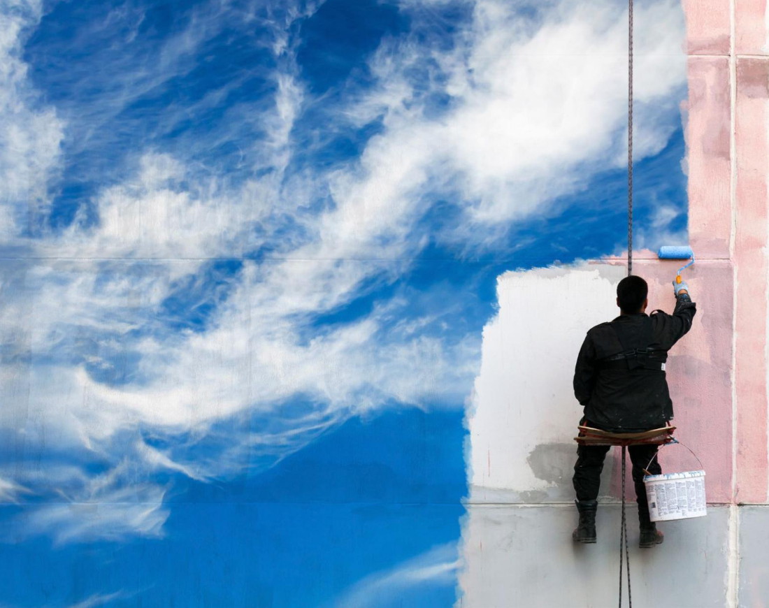 Mann sitzt auf einer Arbeitsbühne und malt einen blauen Himmel auf eine schmutzige Wand