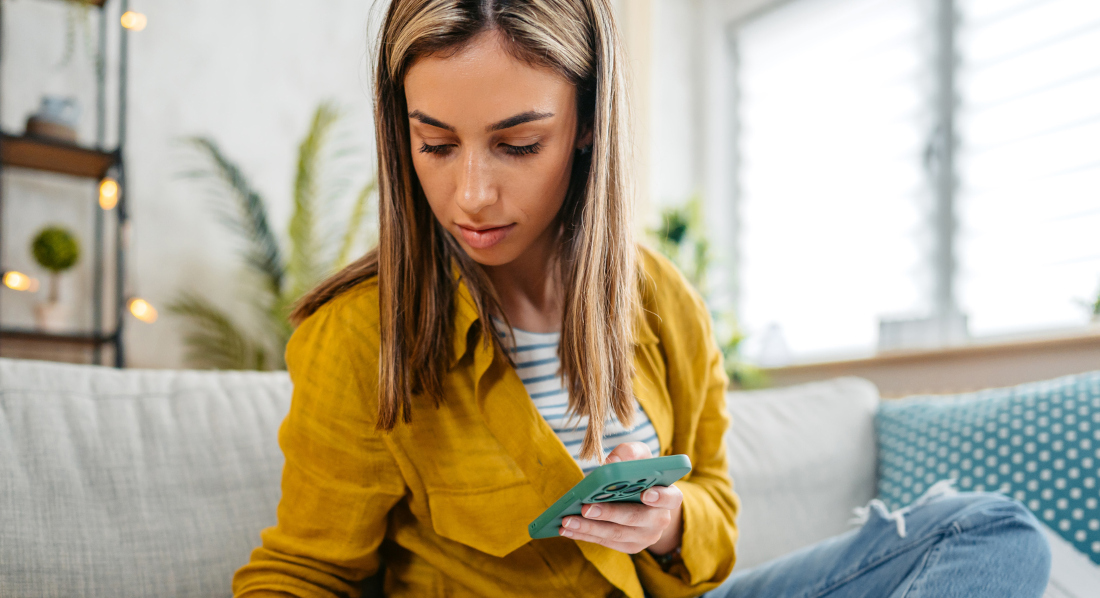 Eine junge Frau sitzt mit ihrem Handy und einigen Blättern Papier auf ihrem Sofa und schreibt etwas auf ein Blatt mit Finanzdaten