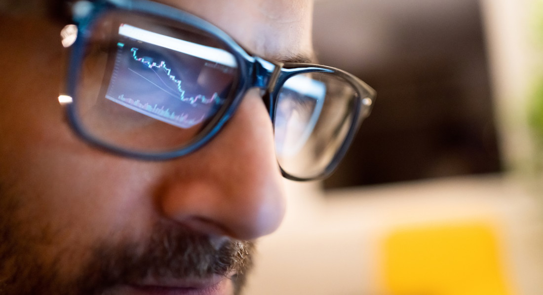 Nahaufnahme vom Gesicht eines bärtigen Mannes, in dessen Brille sich der Kursverlauf einer Aktie spiegelt