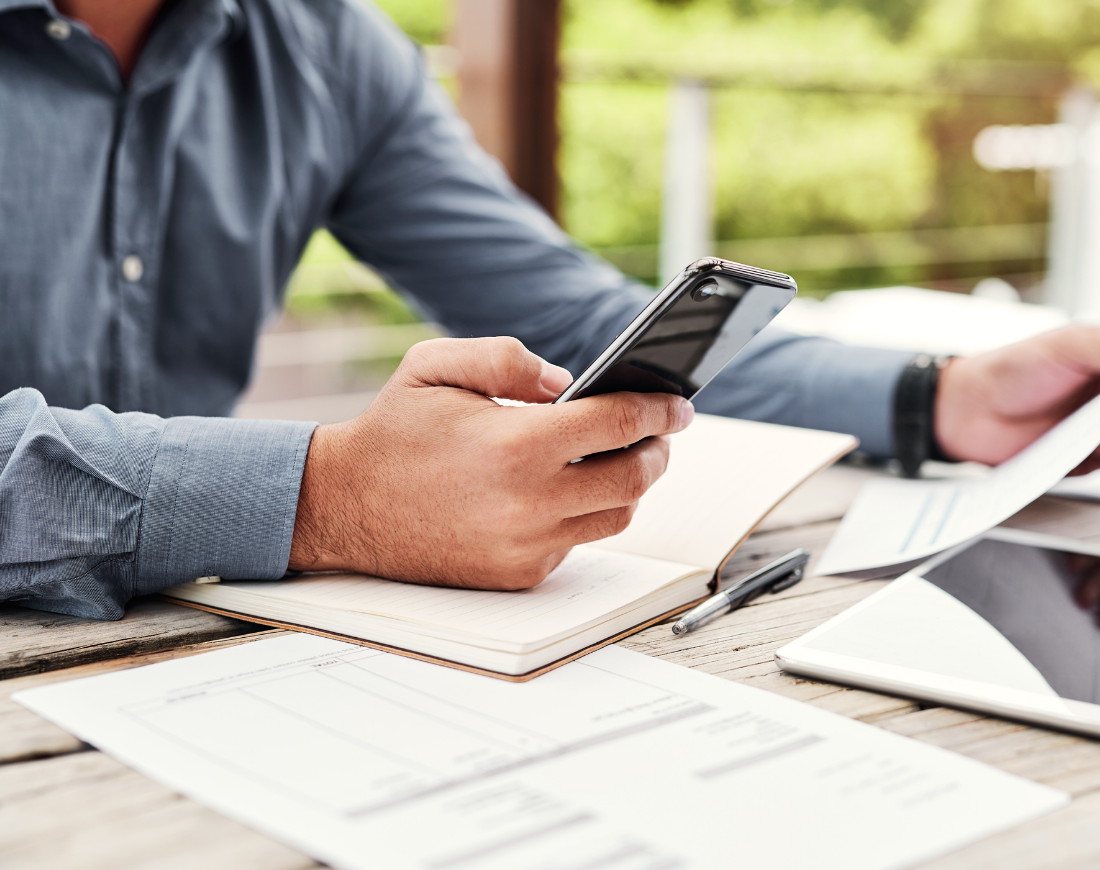 Ein Mann sitzt mit seinem Smartphone, einem Notizbuch, einem Tablet und einigen ausgedruckten Formularen am Tisch