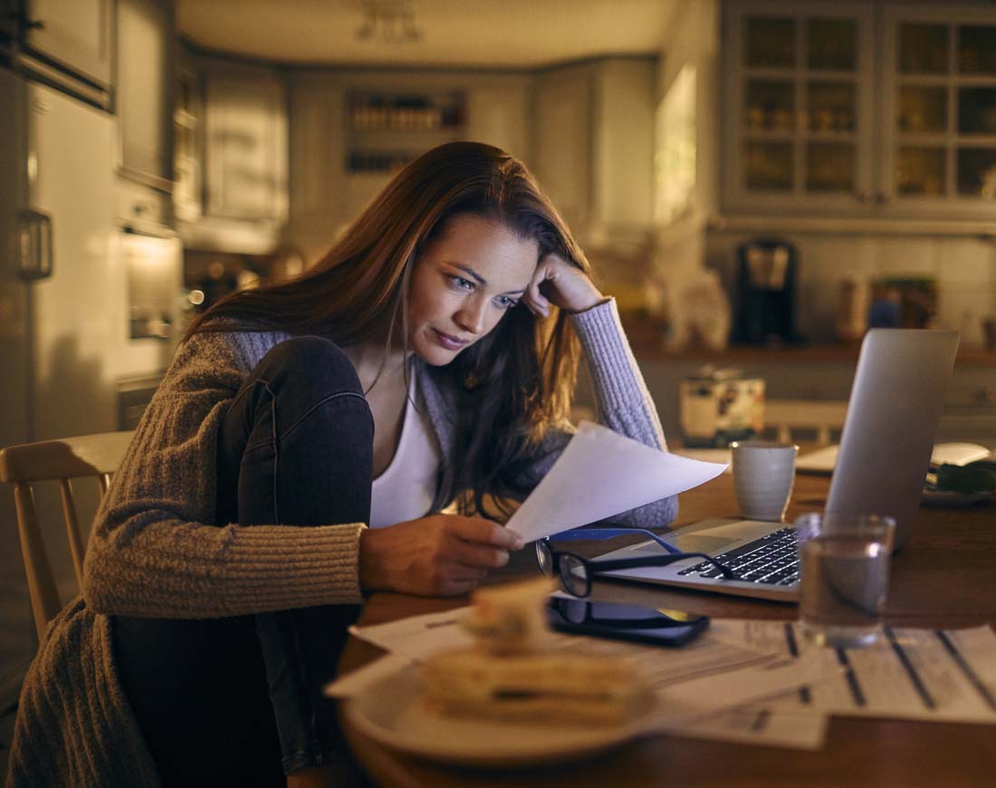 Eine gestresste junge Frau sitzt zu Hause vor ihrem Laptop und blickt verwundert auf Papiere