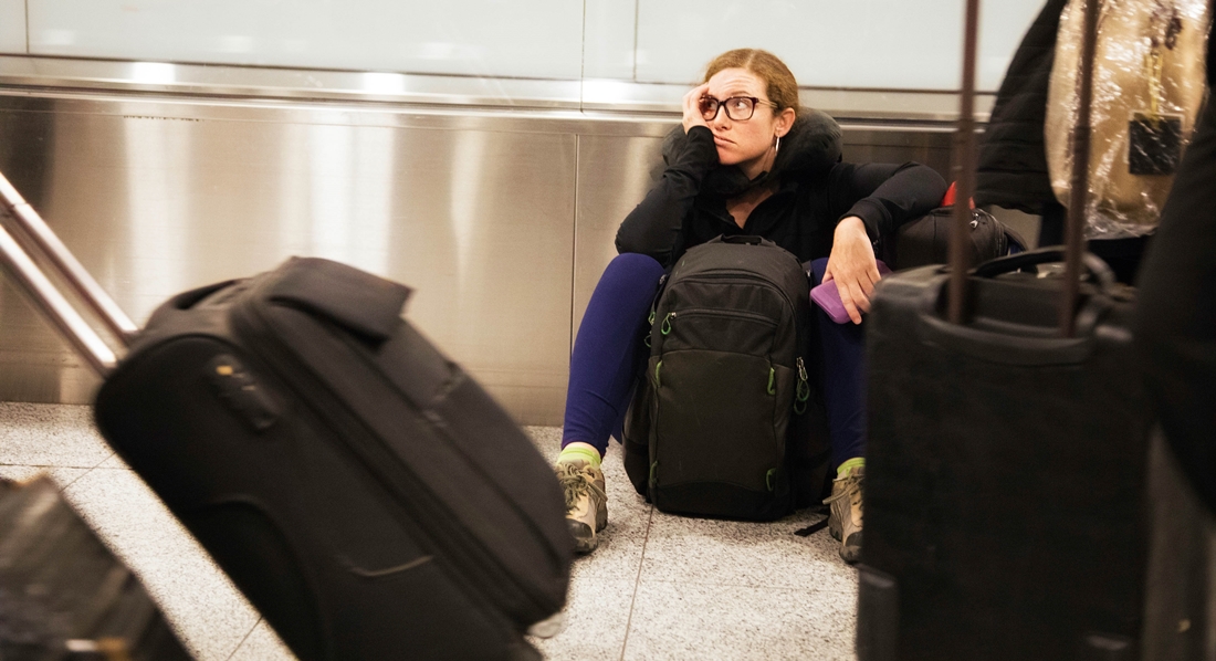 Junge Frau sitzt mit ihrem Rucksack genervt auf dem Boden am Flughafen