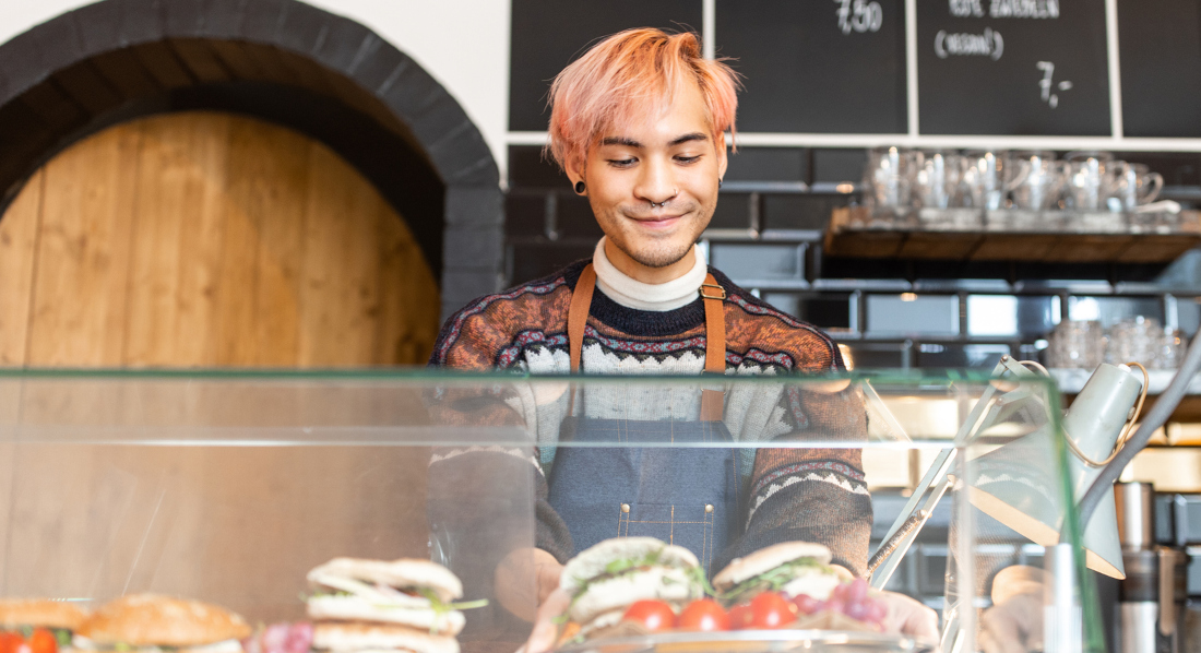 Ein junger Mann mit orange gefärbten Haaren holt einen Teller mit zwei belegten Sandwiches aus Glastheke in einem Café