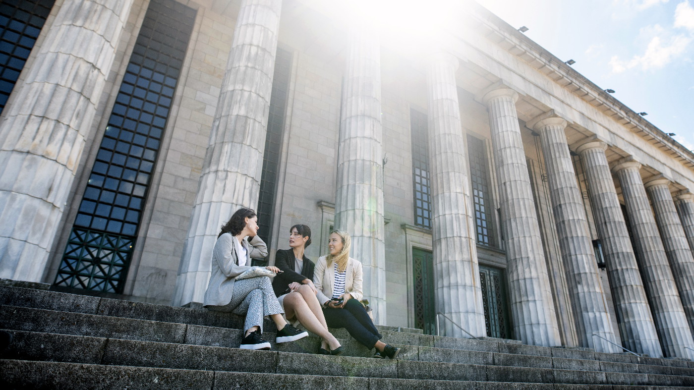 Drei junge Frauen sitzen auf der Treppe vor dem Eingangsportal einer Universität