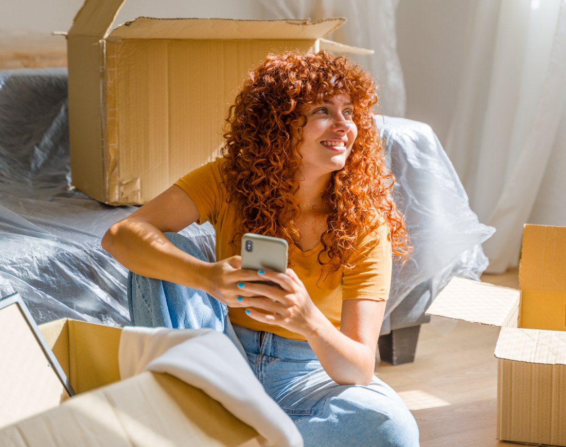 Eine junge Frau sitzt freudestrahlend und mit dem Handy in der Hand zwischen Umzugskartons auf dem Fußboden ihrer neuen Wohnung