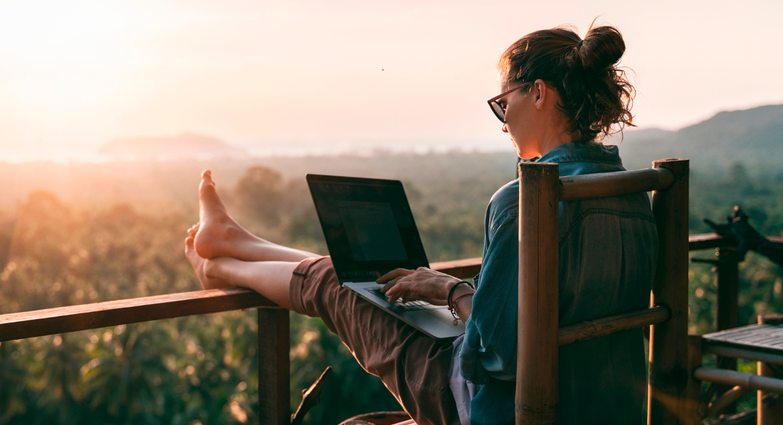 Junge Frau mit Laptop auf dem Schoß arbeitet mit Blick auf tropischen Wald und Meer