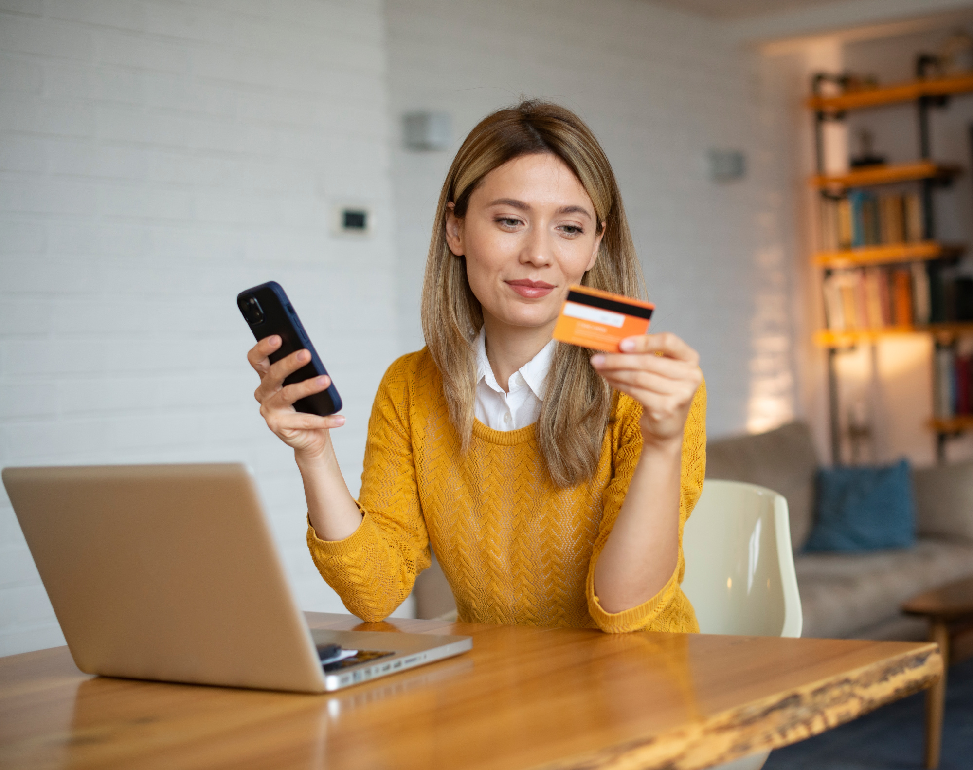 Eine Frau mit Handy und Bankkarte in den Händen führt eine SEPA-Überweisung auf einem Laptop aus