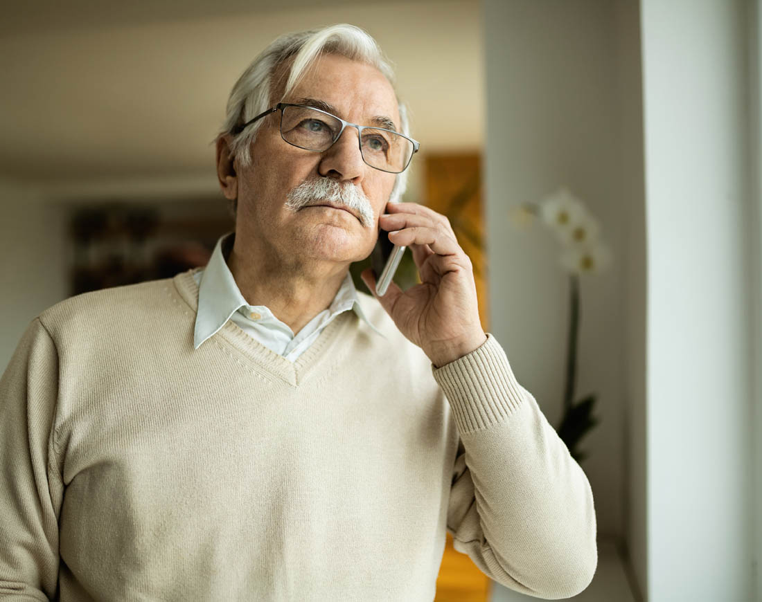 Ein älterer Mann telefoniert zu Hause mit seinem Handy und blickt nachdenklich aus dem Fenster