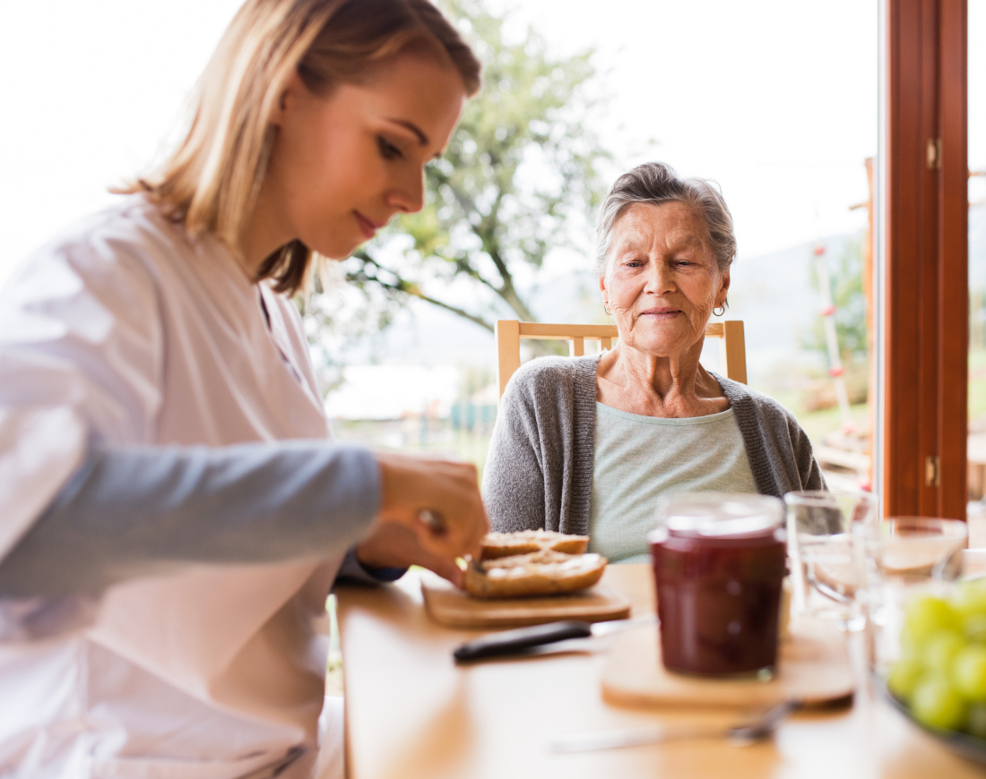 Eine junge Frau schmiert für eine ältere Dame ein Brötchen am Frühstückstischt