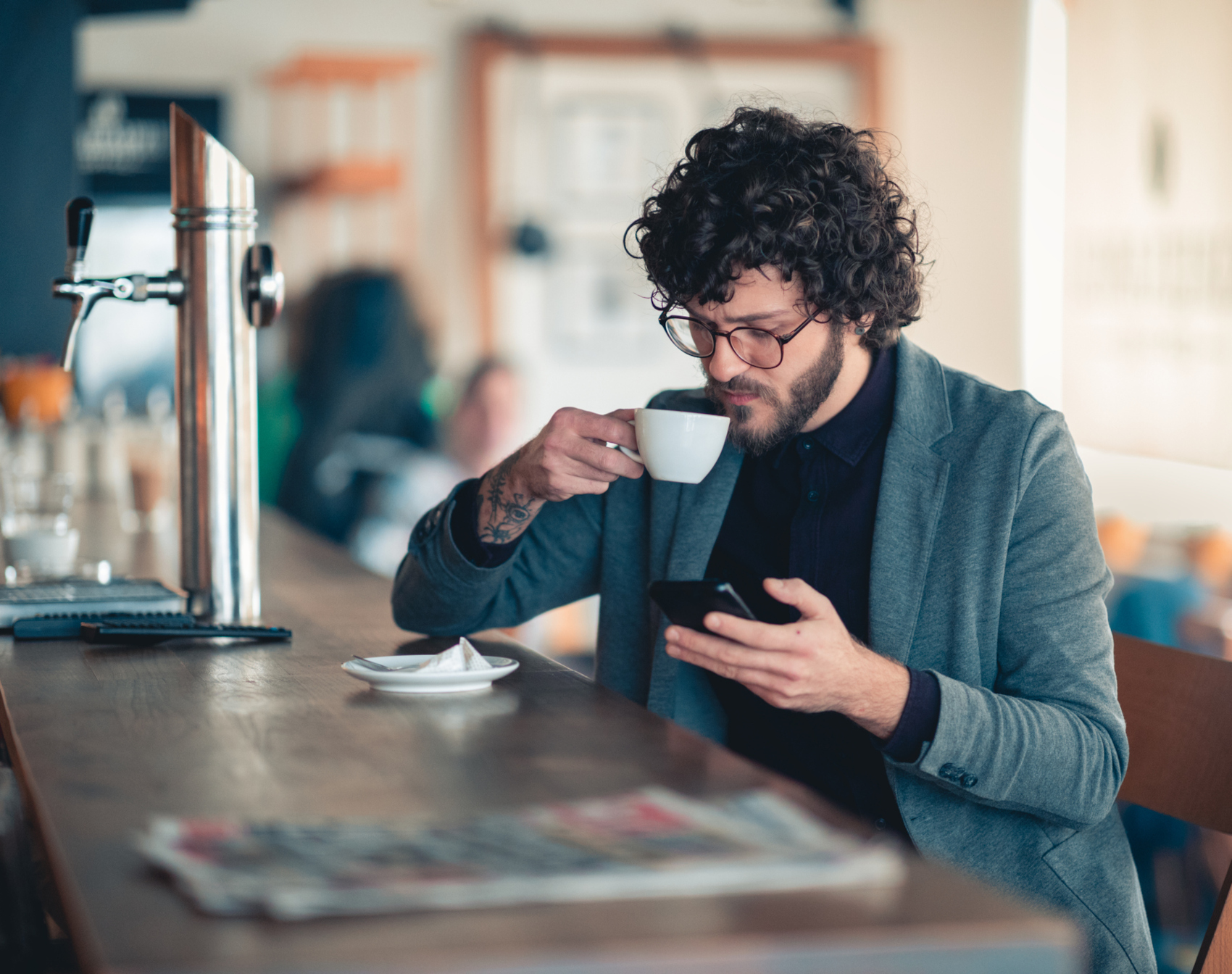 Ein bärtiger Mann sitzt am Tresen, trink Kaffee und schaut auf sein Smartphone