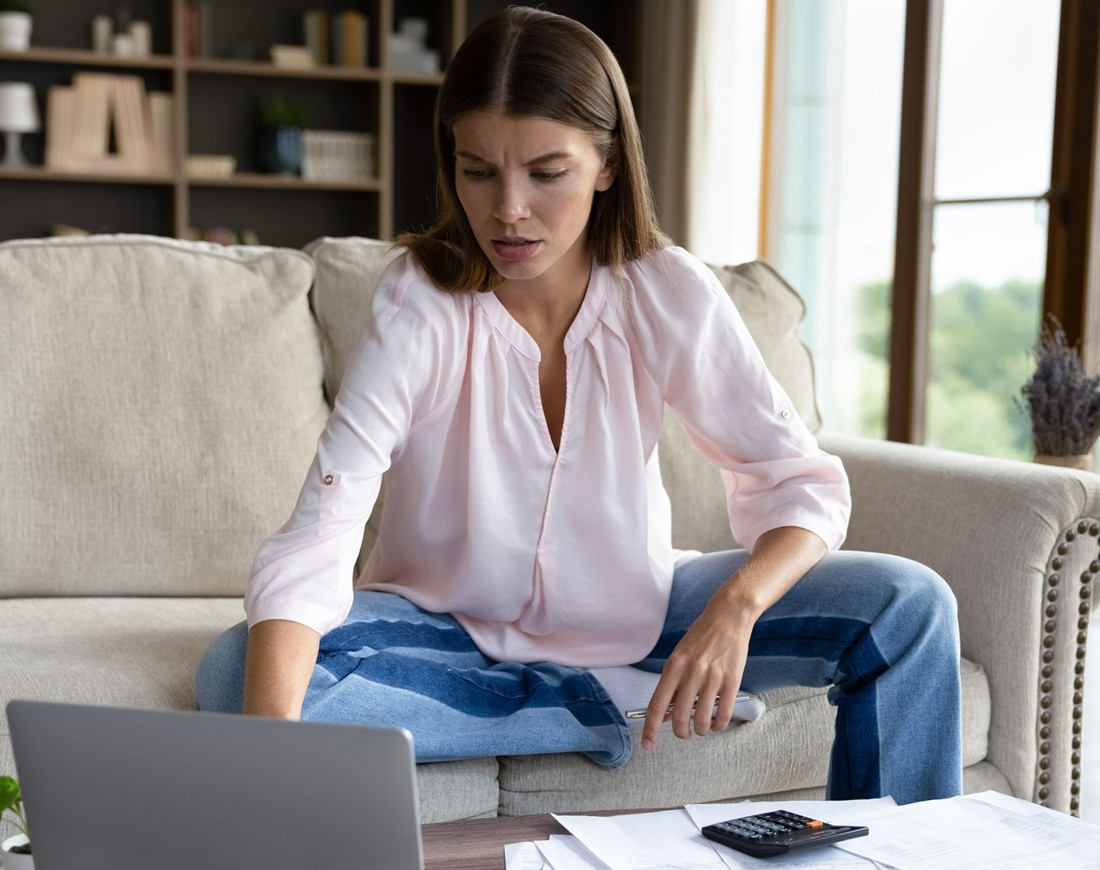 Eine junge Frau hat ihre Rechnungen und einen Taschenrechner vor sich auf dem Tisch und schaut gestresst in einen Laptop
