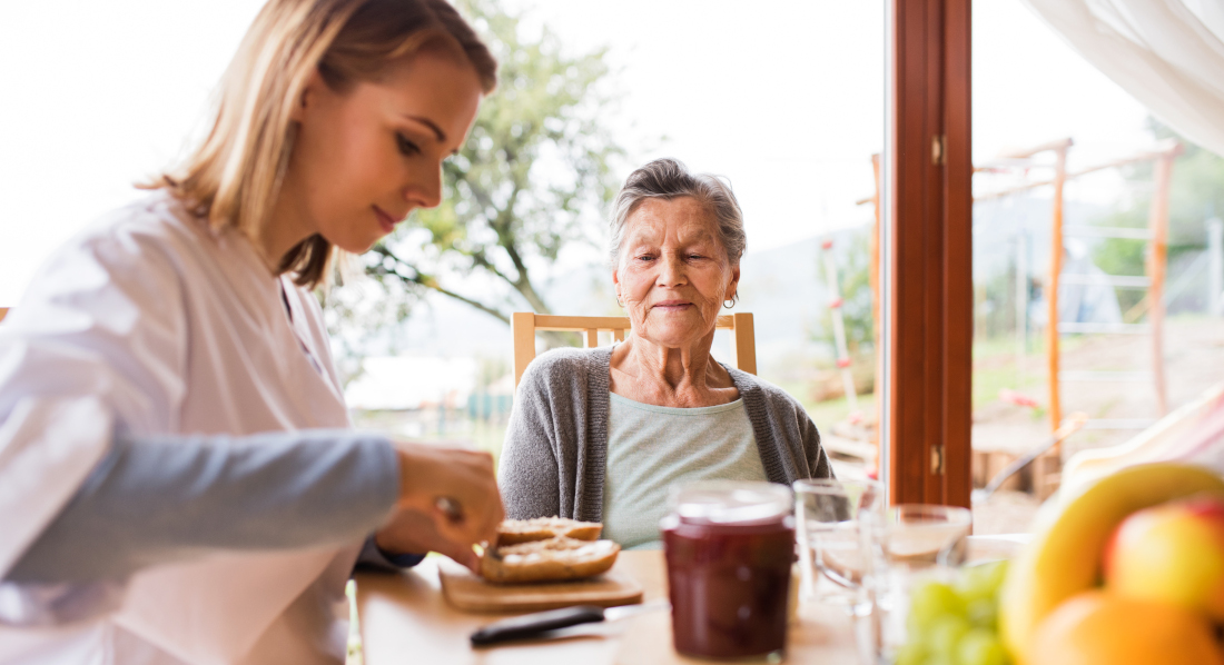 Eine junge Frau schmiert für eine ältere Dame ein Brötchen am Frühstückstischt