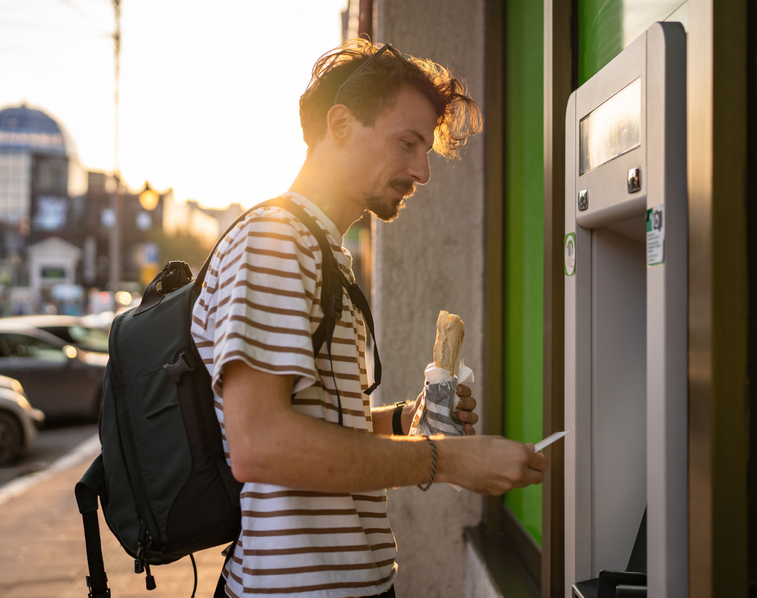Ein Mann beim Geldabheben am Automaten, im Hintergrund sind parkende Autos und Gebäude