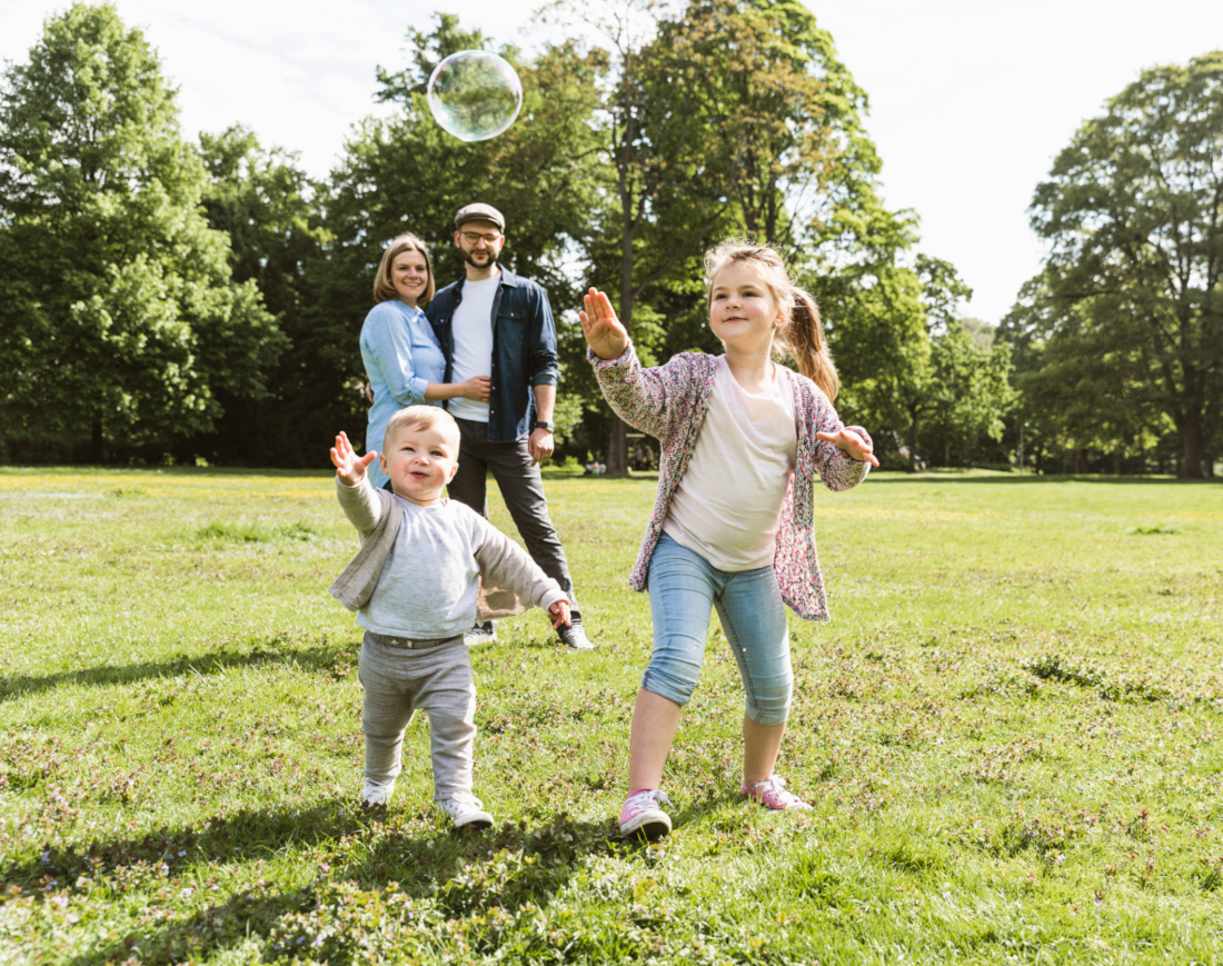 Zwei Kinder laufen hinter einer Seifenblase durch einen Park, hinter ihnen schauen die Eltern zu