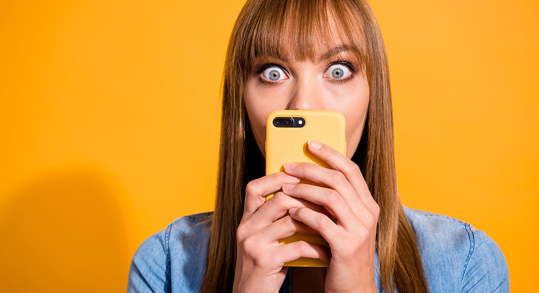 Junge Frau hält sich das Smartphone vor das Gesicht und schaut erstaunt