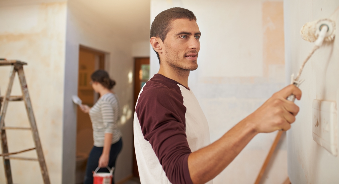 In einer leeren Wohnung führt ein junger Mann einen Farbroller auf einer hellen Wand.