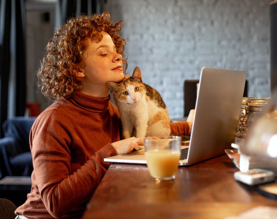 Eine junge Frau arbeitet an ihrem Laptop, während sich ihre Katze an ihr Kinn schmiegt