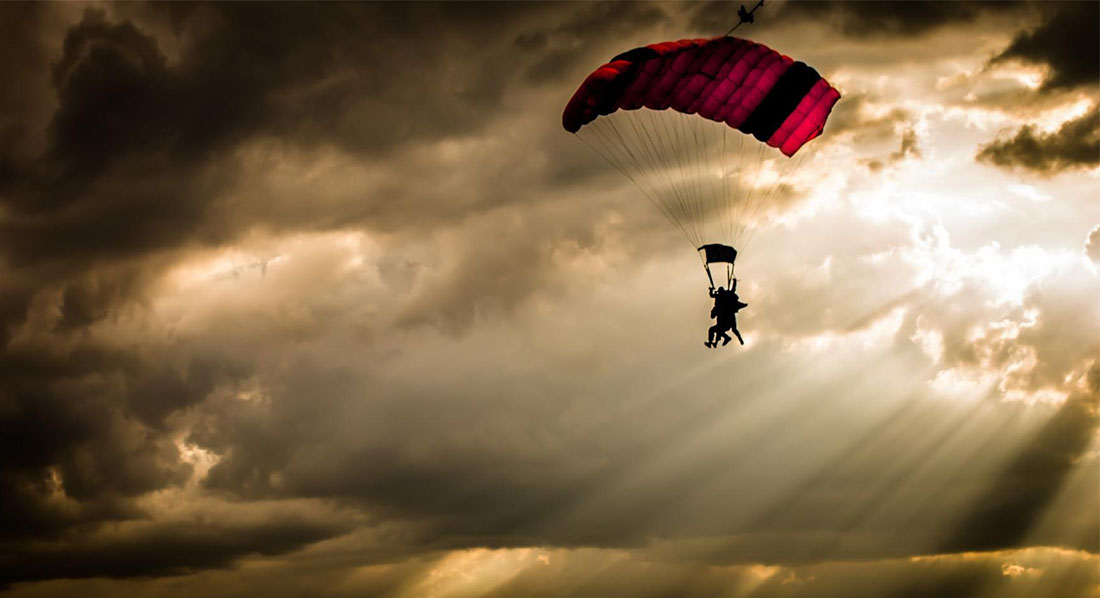 Ein Fallschirm schwebt vor dem Hintergrund düsterer Wolken, durch die einzelne Lichtstrahlen brechen, herab