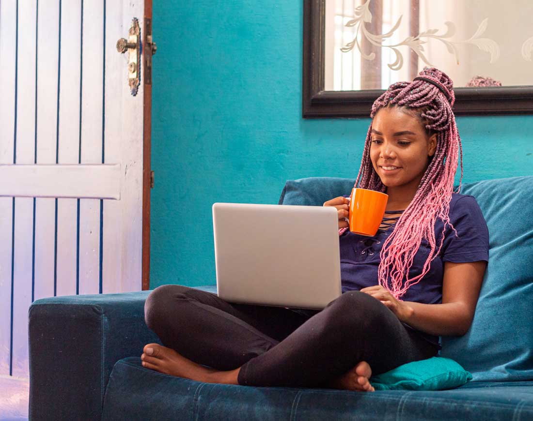 Junge Frau sitzt mit Becher und Laptop auf der Couch