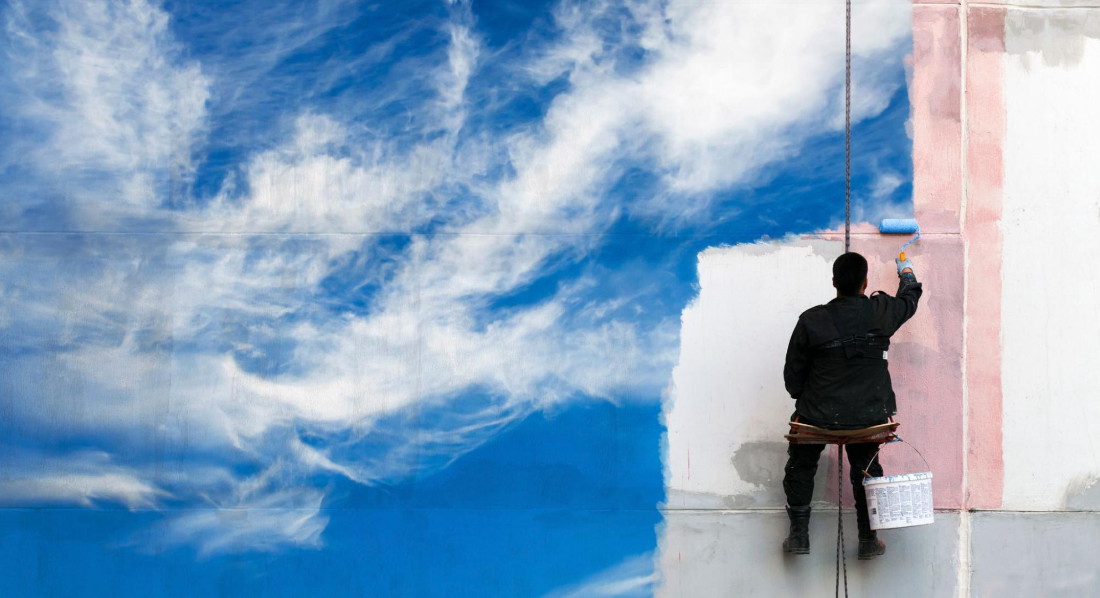 Mann sitzt auf einer Arbeitsbühne und malt einen blauen Himmel auf eine schmutzige Wand