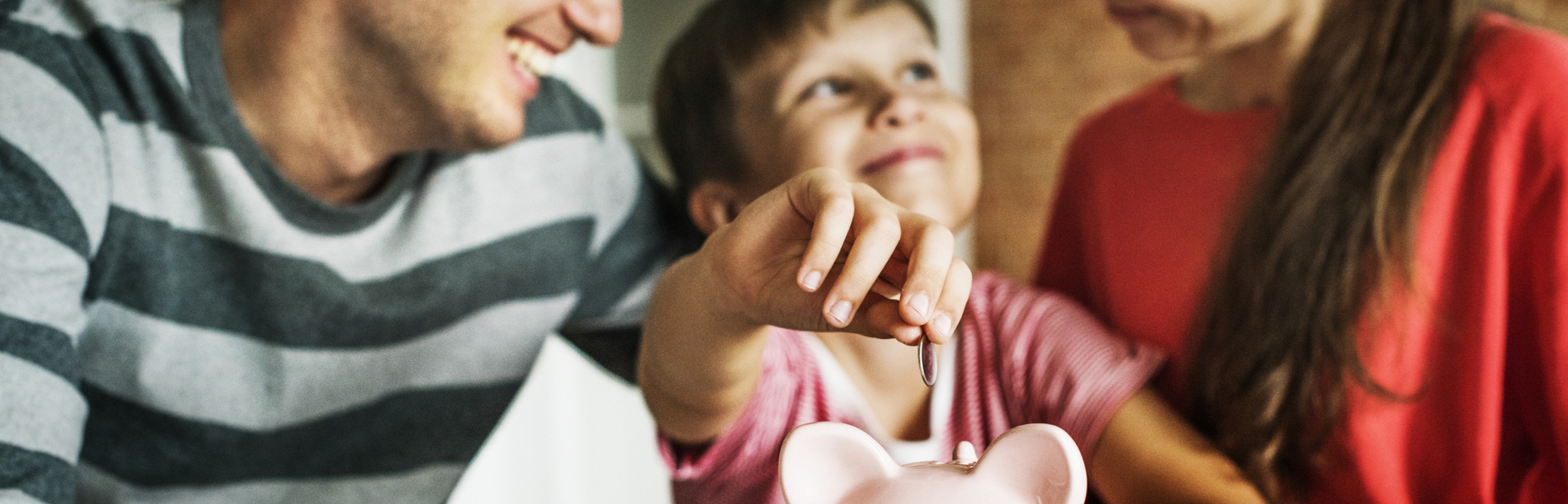 Familie sitzt vor einem Sparschwein, der Sohn wirft ein Geldstück hinein