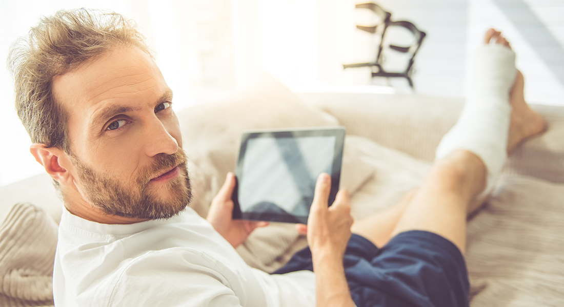 Ein junger Mann liegt mit einem Gipsbein auf einem Sofa und hält ein Tablet in der Hand