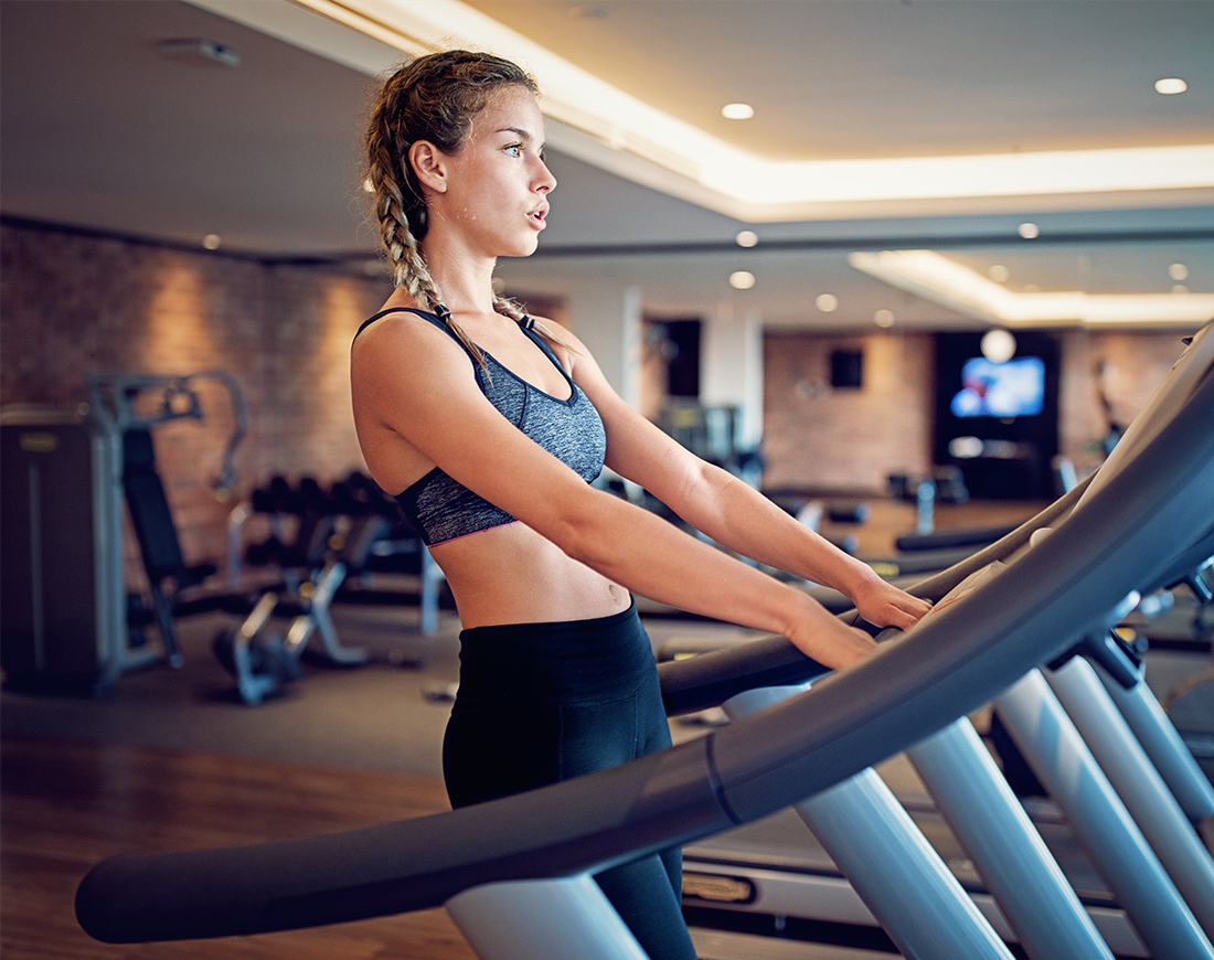 Eine Frau steht alleine auf dem Laufband in einem sonst leeren Fitnessstudio und atmet durch