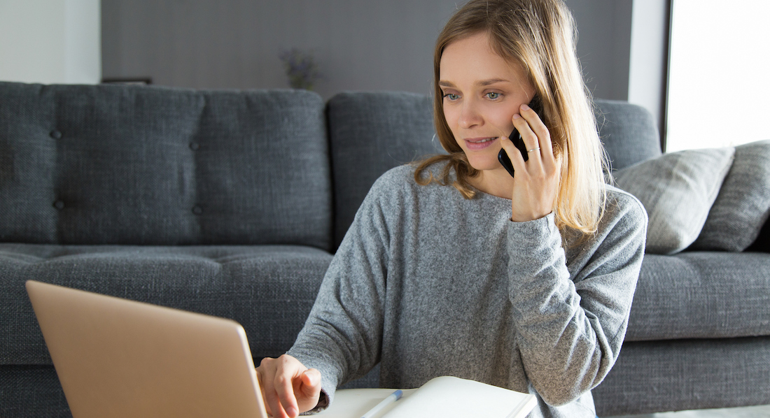 Eine junge Frau sitzt vor Laptop und Notizbuch und telefoniert mit ihrem Smartphone, um ihre Kreditkarte zu sperren.