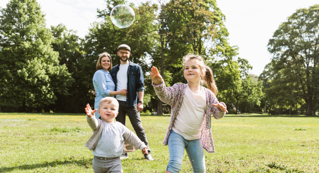 Zwei Kinder laufen hinter einer Seifenblase durch einen Park, hinter ihnen schauen die Eltern zu