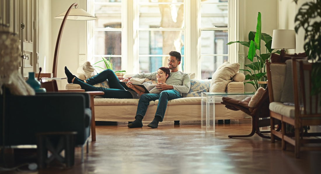Hausratversicherung: Glücklich wirkendes junges Paar auf einem hellen Sofa in einer geschmackvoll möblierten Altbauwohnung beim Betrachten eines Buches