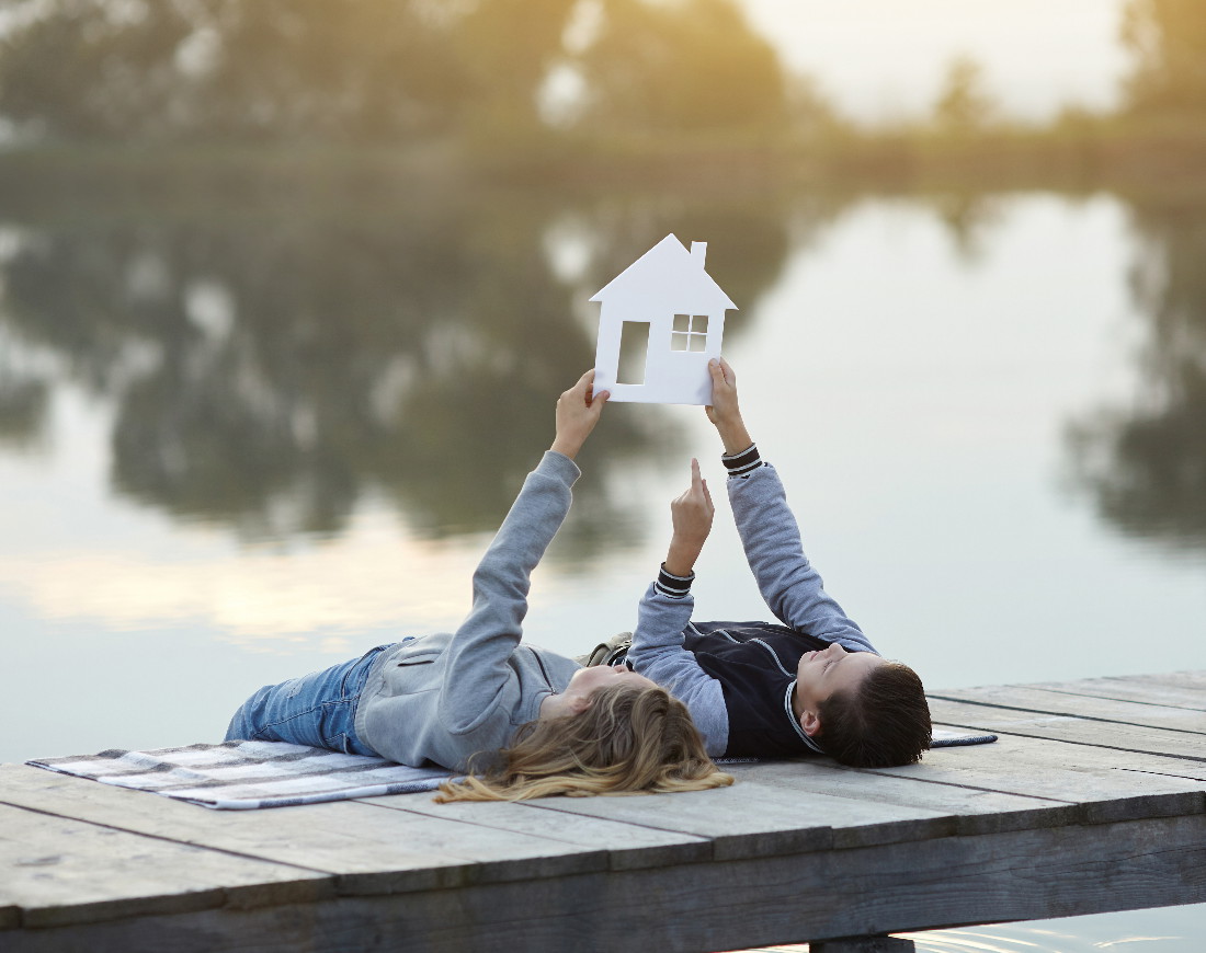 Ein Paar liegt auf dem Rücken auf einem Steg am See und hält ein Haus aus Papier in die Luft