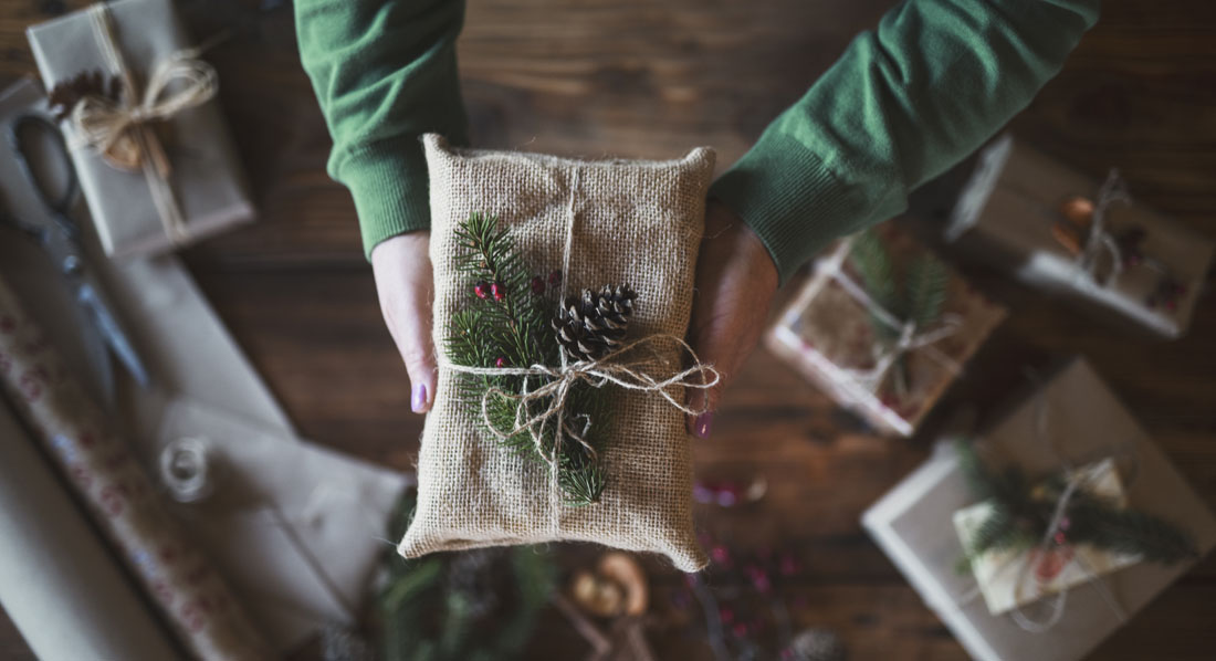 Eine Frau hält über einem Basteltisch eine Geschenkverpackung aus Stoff und mit Tannengrün verziert hoch